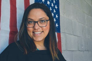 Image: Woman with dark hair and glasses smiling in front of an American flag